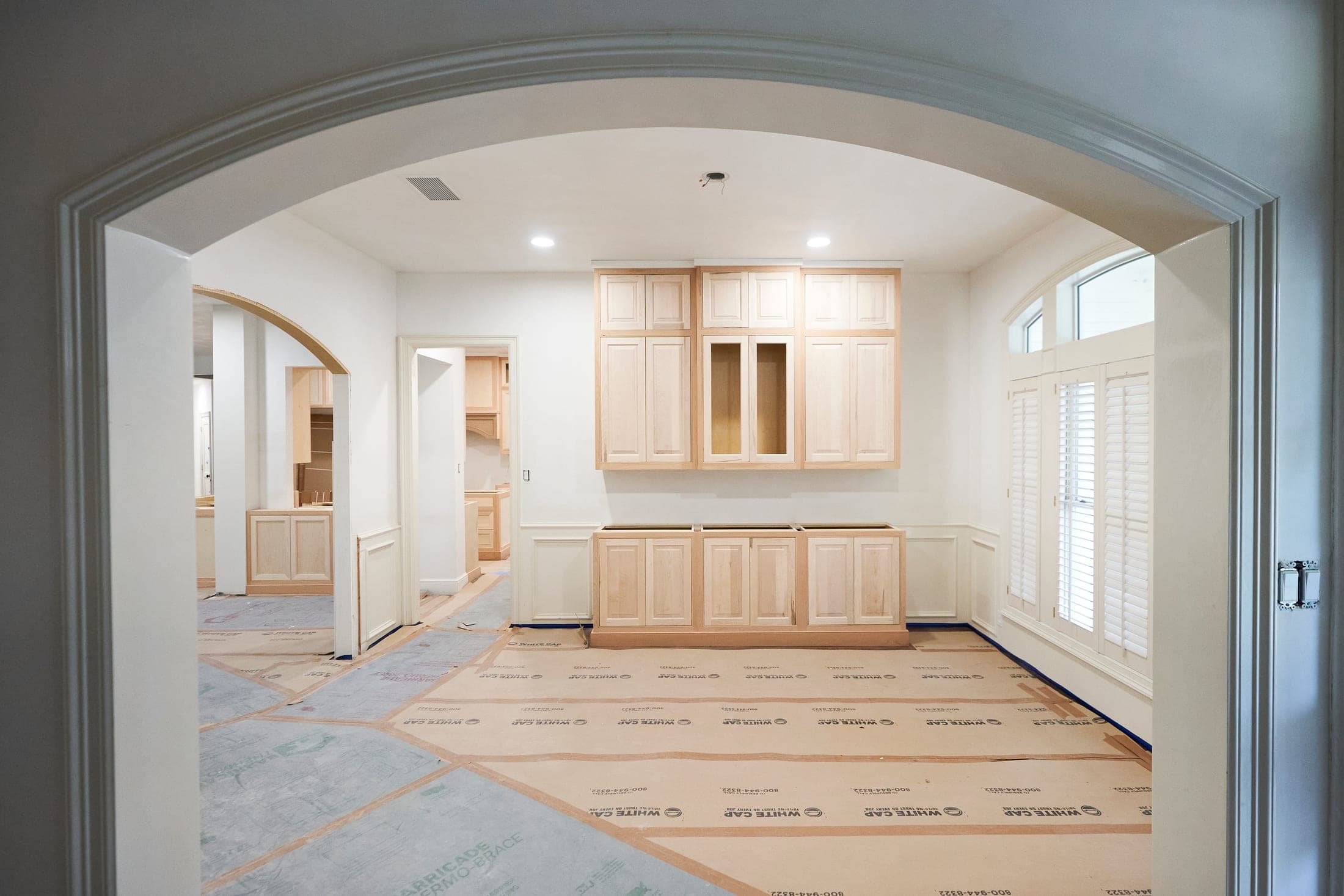 Unfinished cabinetry installed along a kitchen wall, seen through an arched opening, the floor still under protection