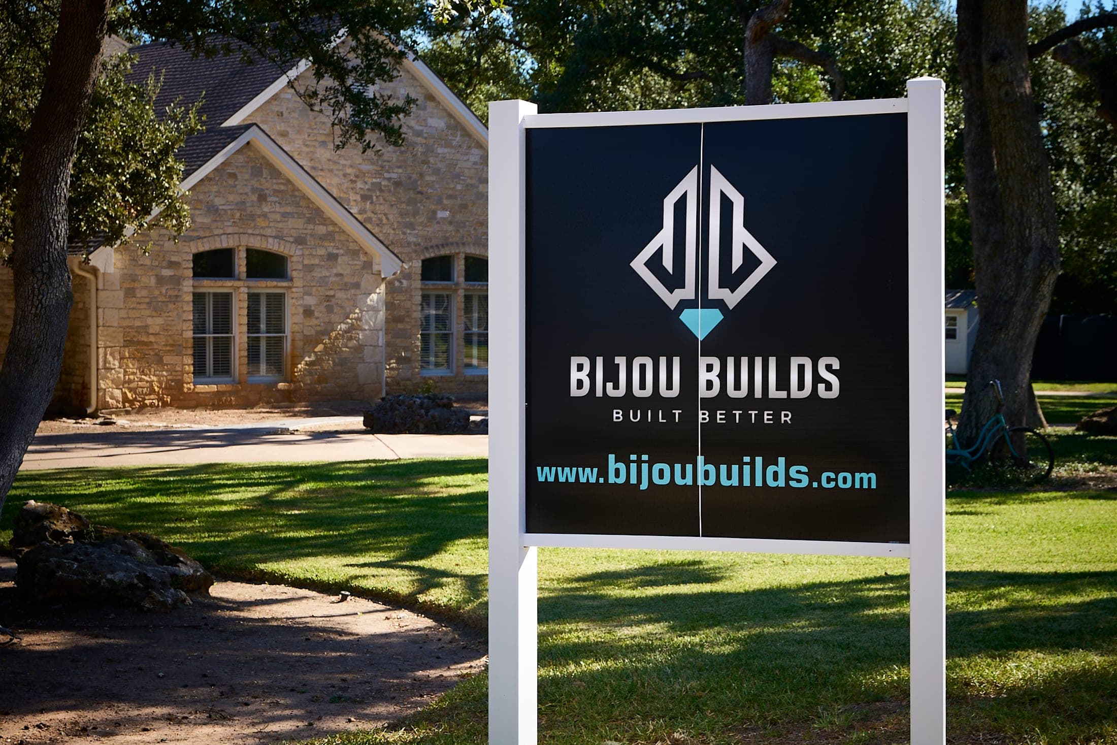 A Bijou Builds yard sign in front of a stone house under live oaks