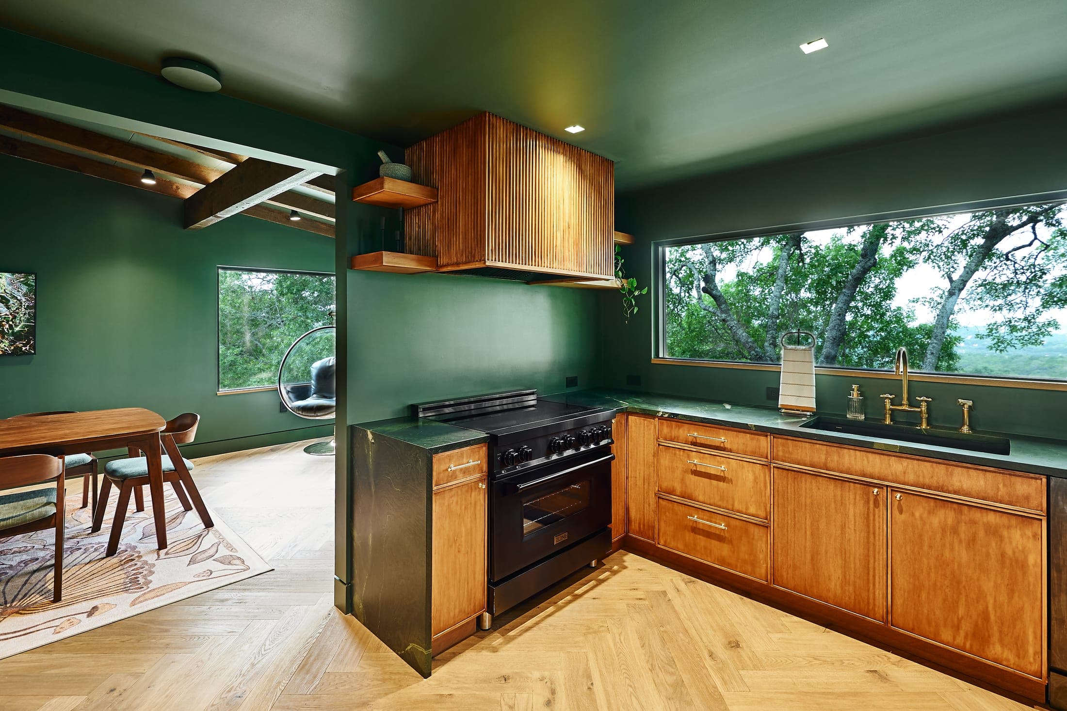 A kitchen with deep green walls, warm wood cabinetry, a black range and a long picture window onto the trees