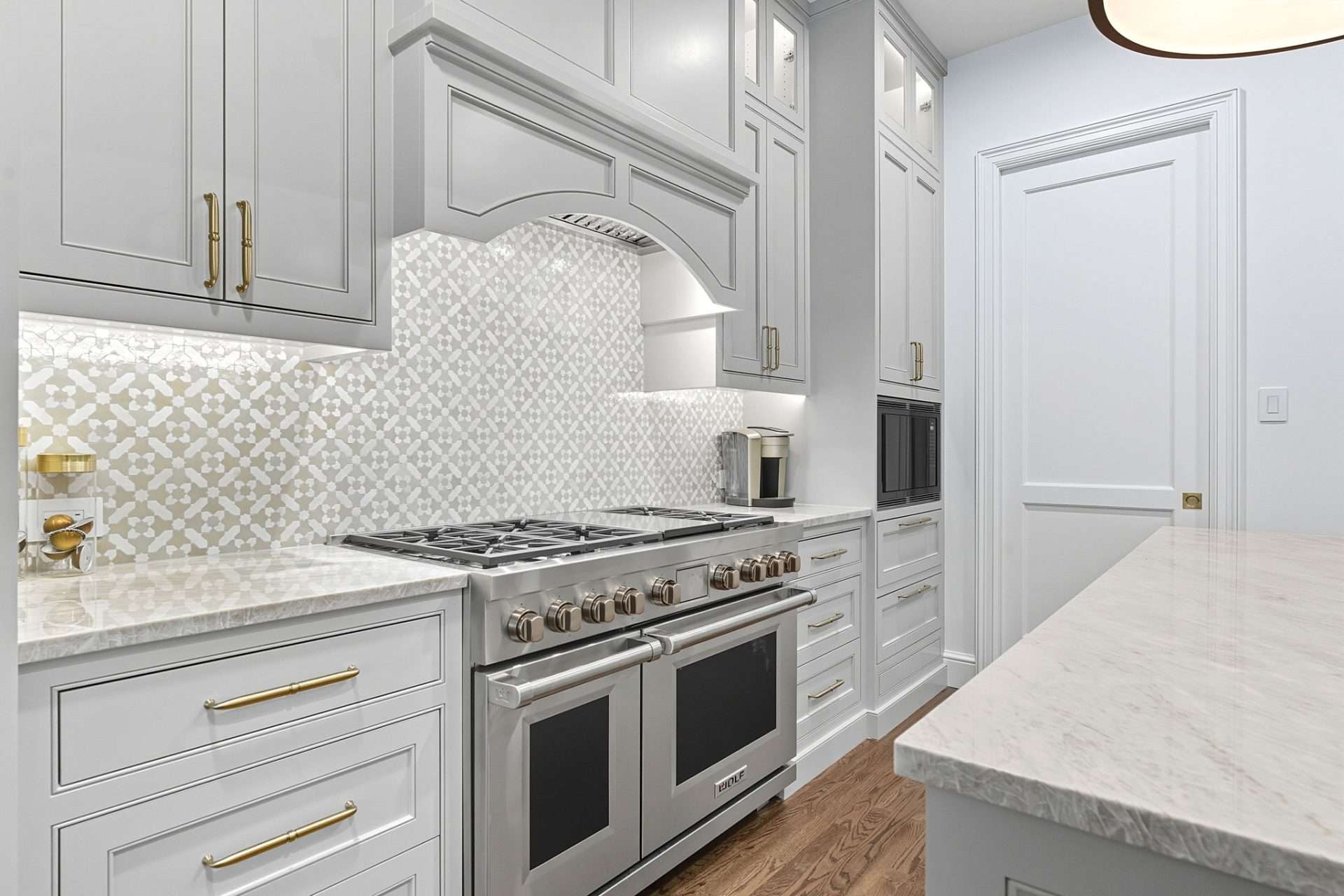 A kitchen with pale grey inset cabinetry, brass pulls, a patterned tile backsplash, a six-burner range with double ovens and marble-look counters